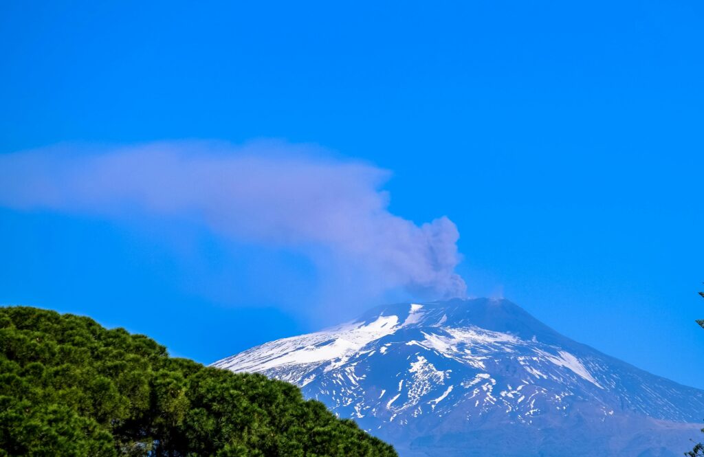 A scenic view of a snow-covered active Mount Etna volcano with smoke under a bright blue sky.